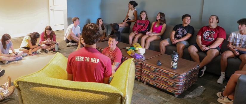 Students sit in an Honors Residence lounge as an RA with his back to us begins their first floor meeting