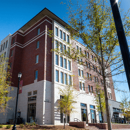 Students walking and on bikes walk on a brick courtyard.  Behind them is a six story brick residence hall flanked by trees.