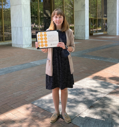 Riley Sutherland stands in front of Thomas Cooper with an award wearing a black and white polka dotted dress and beige cardigan