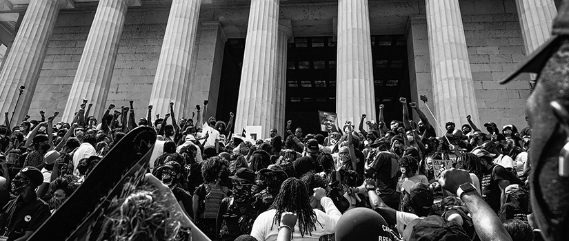 Black and white photograph of a large crowd of protesters gathered on the steps of a columned building, many with raised fists, documenting a demonstration during the summer of 2020.