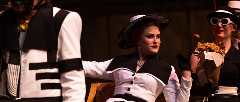 Student performer in black-and-white costume holding flowers while seated on stage with other costumed actors nearby.