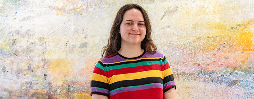 Emily Moffitt stands smiling in front of a textured, abstract mural. She wears a colorful striped shirt and looks confidently at the camera.