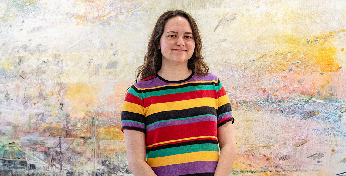 Emily Moffitt stands smiling in front of a textured, abstract mural. She wears a colorful striped shirt and looks confidently at the camera.
