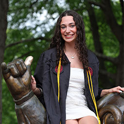 Graduate wearing cap and gown with honor cords, sitting on a statue hand and smiling outdoors.