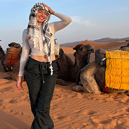 Graduate standing in the desert, smiling with camels in the background.