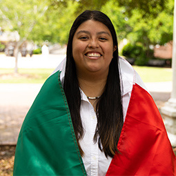 Graduate smiling while draped in a Mexican flag, standing outdoors between columns on a sunny day.