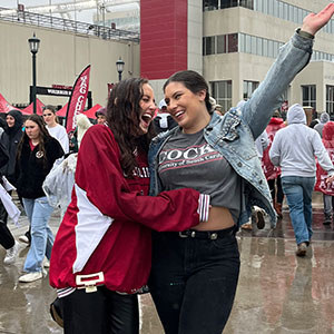 Two students laughing and posing together in the rain at a spirited USC event, surrounded by a crowd.