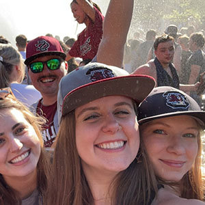 Students smiling and wearing University of South Carolina Gamecock hats, enjoying a fountain celebration with a cheering crowd.