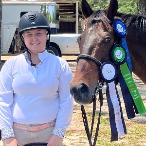 Student wearing equestrian gear stands beside a horse adorned with multiple award ribbons after a competition at the Palmetto Classic.