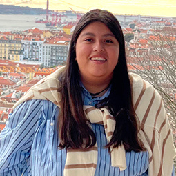 Student smiling in front of a scenic cityscape with red rooftops and a large suspension bridge in the background.