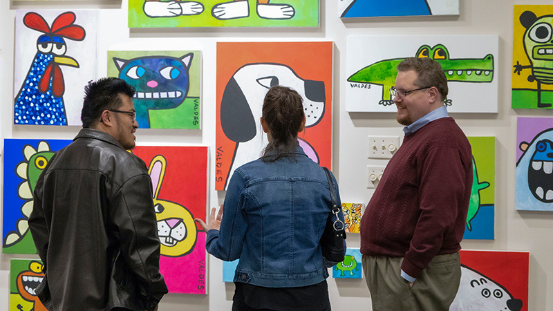 Three people stand in front of a brightly colored gallery wall filled with playful, cartoon-style animal paintings by artist Marius Valdes, engaging in conversation while viewing the artwork.