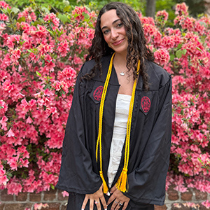 Graduate in a black USC cap and gown with honor cords poses in front of blooming pink flowers, smiling on a spring day.