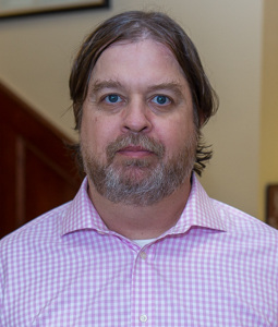 Jeffrey wears a pink and white checked oxford shirt and smiles at the camera