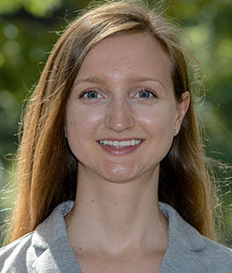 Portrait of a woman with long straight blonde hair, wearing a grey blazer, smiling outdoors with green trees behind her.
