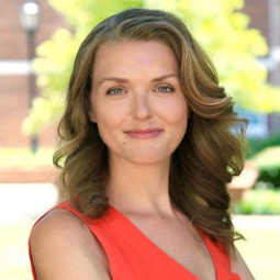 Jennifer wears a red dress and is smiling, she stands against an outdoor background