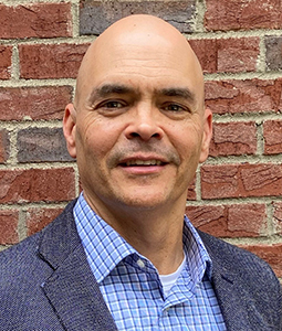 a man smiling in front a brick wall with a blue blazer on a checked button up shirt