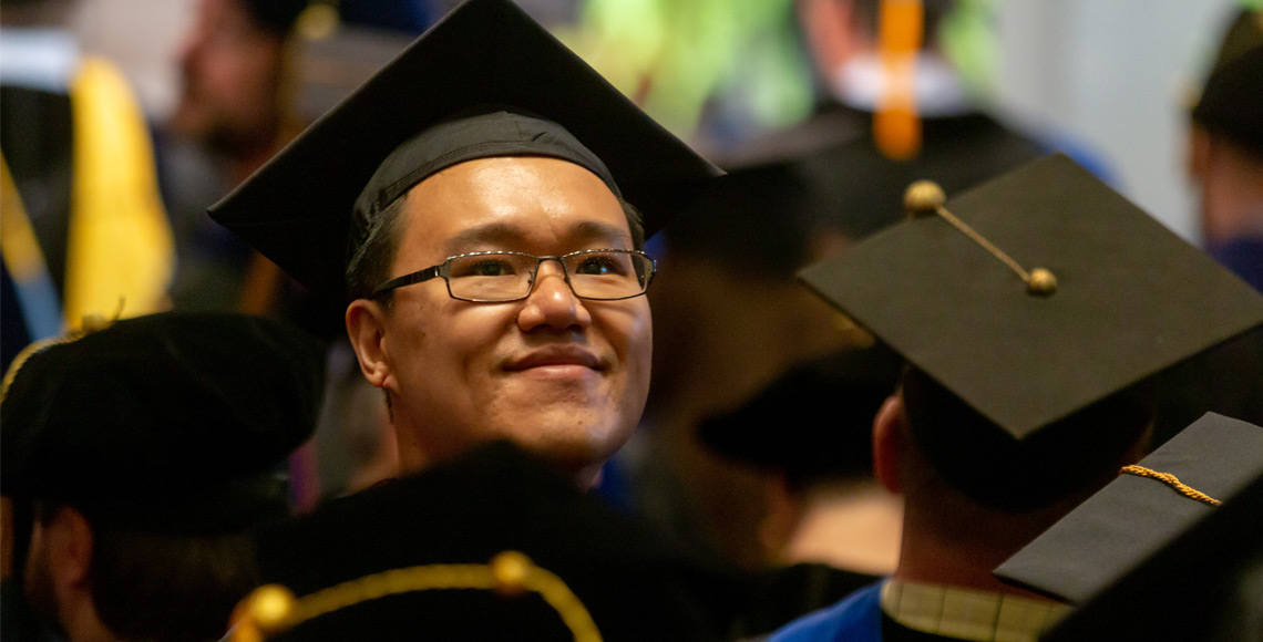 Graduate student wearing a commencement cap smiles at the camera while other students in commencement caps stand around him facing away.