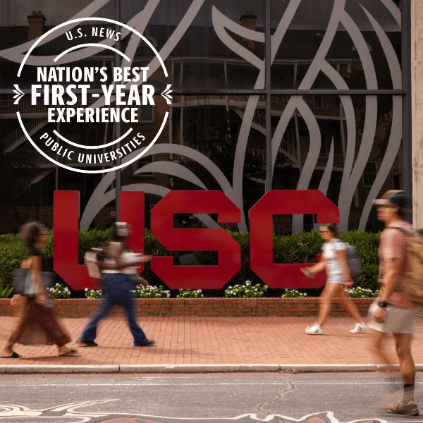 University of South Carolina (USC) students walk past a large USC logo outside a campus building. A graphic reads, “U.S. News Nation’s Best First-Year Experience — Public Universities.”