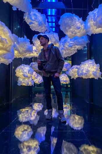 A man in casual clothing stands in front of a bckground of clouds