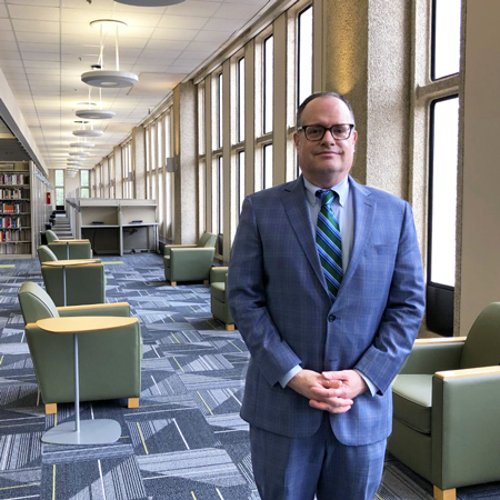 david banush stands in a library with rows of bookshelves on one side and large windows on the other