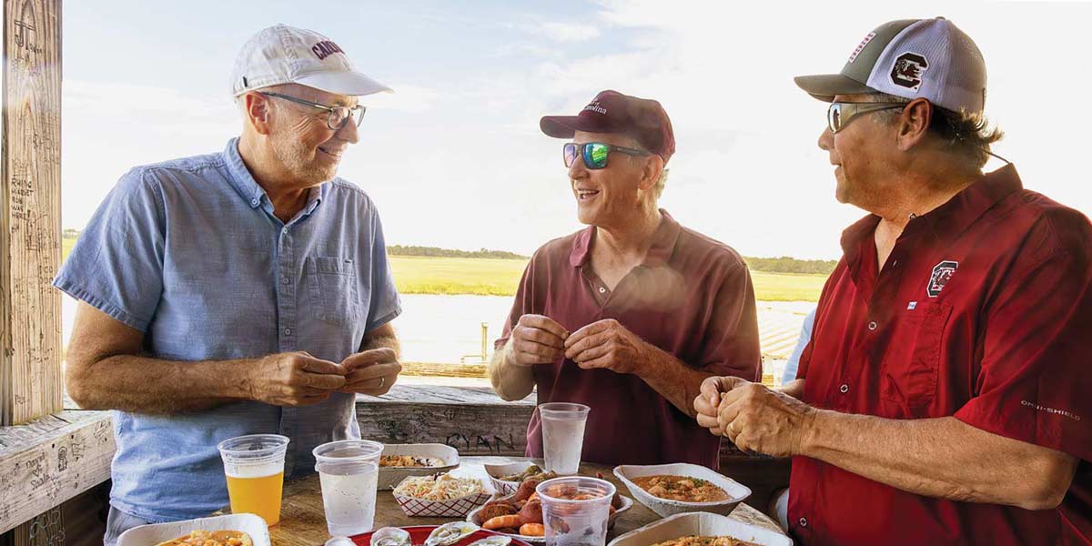 three men wearing ball caps sit on a deck eating